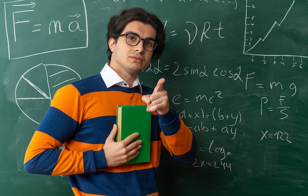 serious young caucasian geometry teacher wearing glasses standing front chalkboard classroom holding book 141793 106363